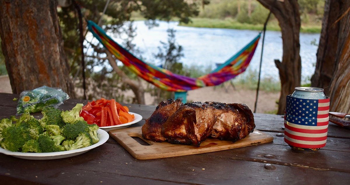 Roast chicken, chopped bell peppers, and broccoli on plates with a beer on a picnic table with a hammock and river in the background