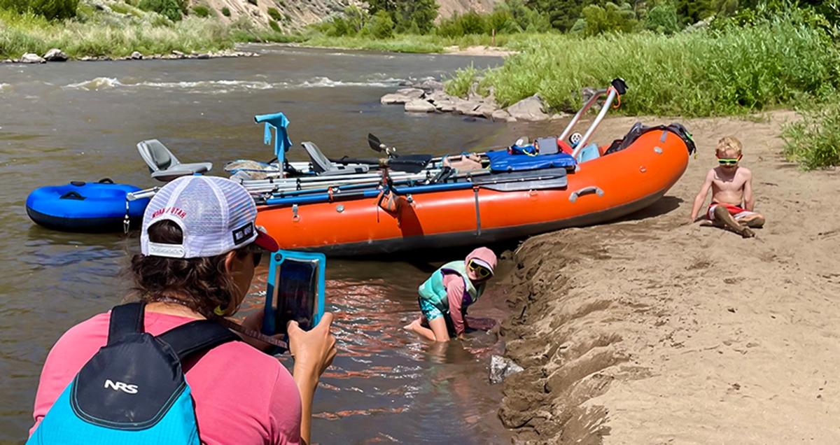 A mom taking photos of her kids on a riverbank with her phone in a waterproof case. There is a raft on the bank and the river running in the background