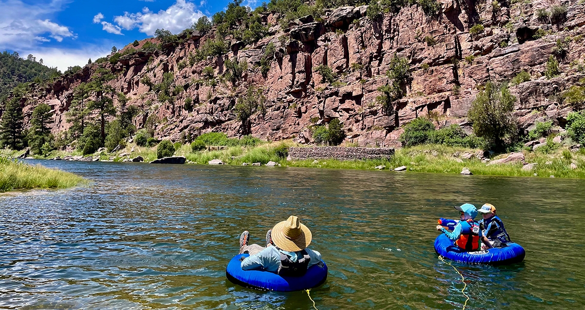 A man with a straw hat floating on a blue inner tube next to two kids on another blue inner tube with a river and tree covered canyon walls in the background.