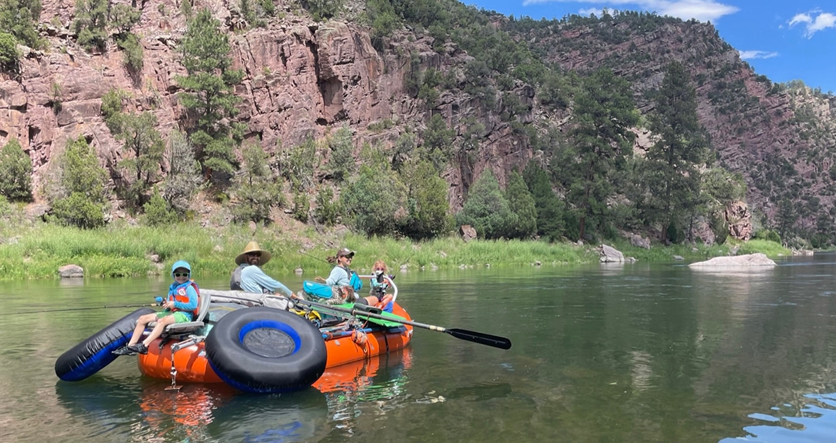 Family floating down river on a river raft with oars, red cliffs with green bushes in the background and on the banks