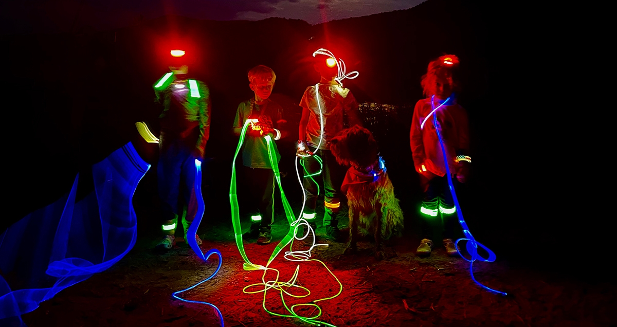 Four kids on a riverbank at night covered with multicolored light up rope, headlamps, and other gear that lights up.